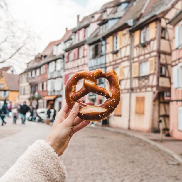A hand holding a salted pretzel on a street with colorful traditional houses in the background