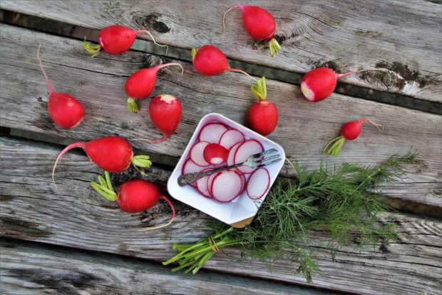 Fresh radish and sliced radish arranged on a wooden surface with green herbs and a small fork