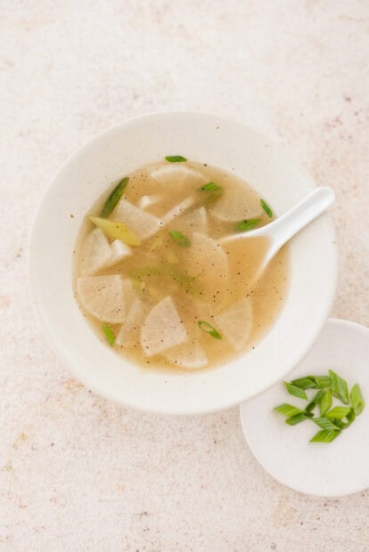 A bowl of radish soup garnished with chopped green onions and black pepper in a white bowl
