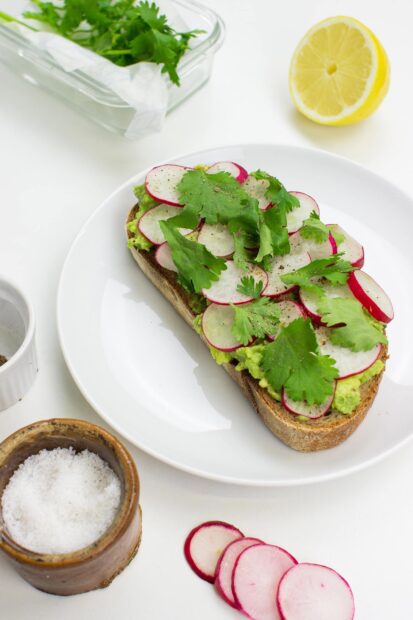 A slice of bread topped with sliced radish and fresh herbs on a white plate