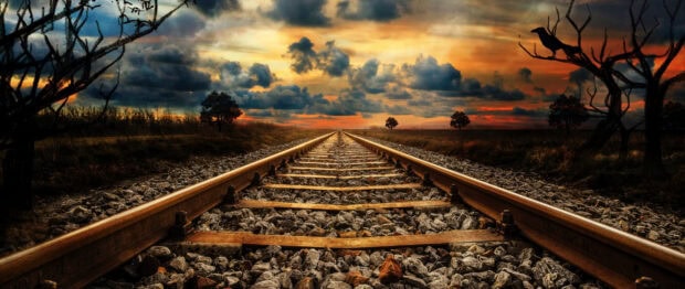 Railway tracks extending into the horizon under a dramatic sunset sky with clouds and silhouetted trees