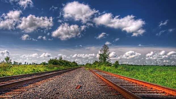 Railway tracks stretching through green fields under a cloudy blue sky in a rural landscape