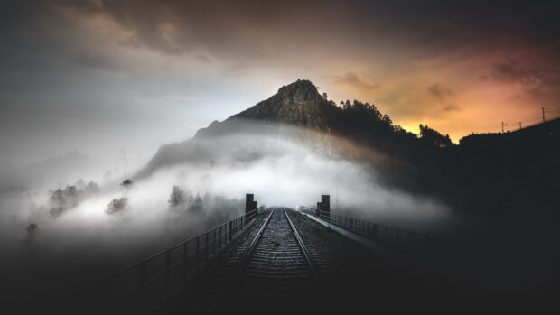 Railway rail tracks leading towards a misty mountain under a dramatic sunset sky