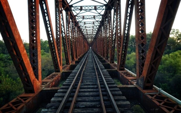 A rusted railway bridge structure with tracks extending through a green forest landscape