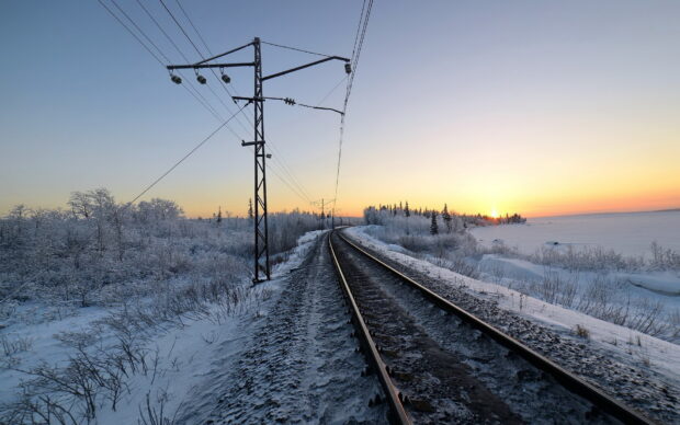 Snow covered railway track stretching through a winter landscape at sunrise with trees and electric poles