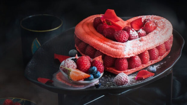 A heart shaped dessert filled with fresh raspberries and decorated with rose petals on a black plate
