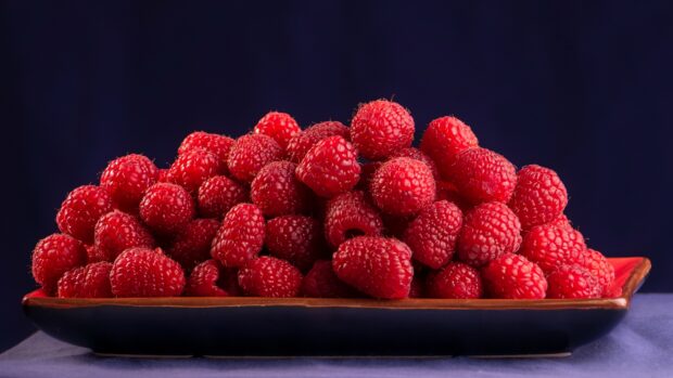 A close up view of fresh red raspberry on a ceramic plate against dark background