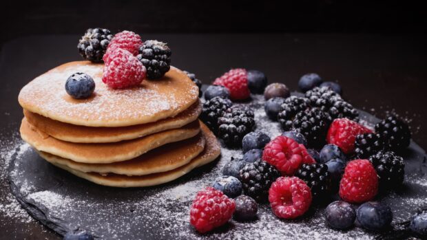 A stack of pancakes topped with fresh raspberries surrounded by blueberries and blackberries on a dark surface