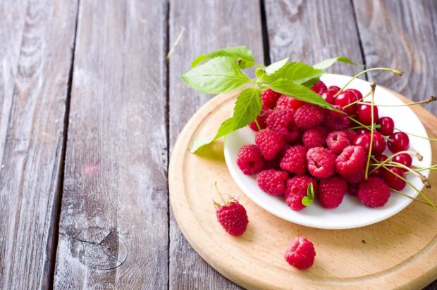 Fresh raspberry fruit with green leaves on a white plate placed on a wooden surface