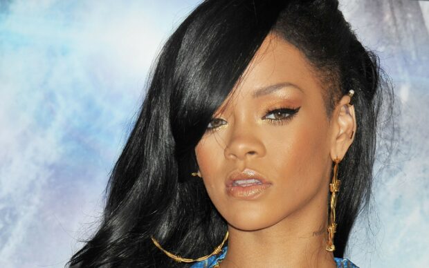 Rihanna with long black hair and golden earrings posing against a blue background