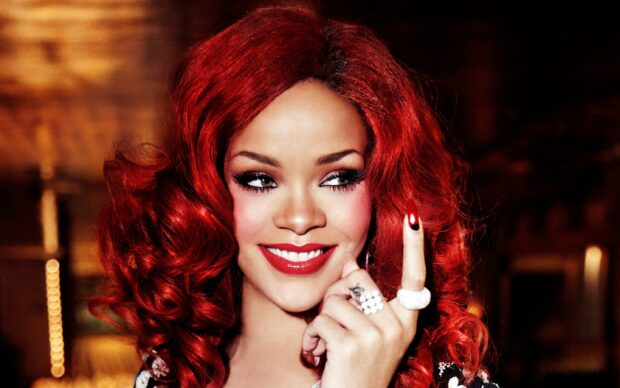 Rihanna with red hair smiling and showing a raised finger with rings in a close up portrait