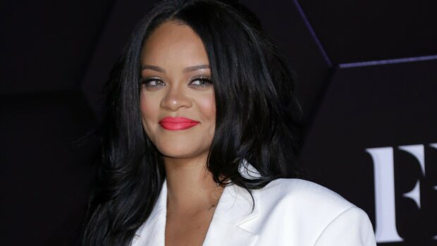 Rihanna with long black hair and red lipstick smiling in a white blazer on a dark background