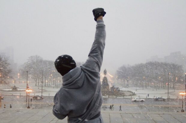 Man in gray hoodie raising fist on snowy steps inspired by Rocky theme