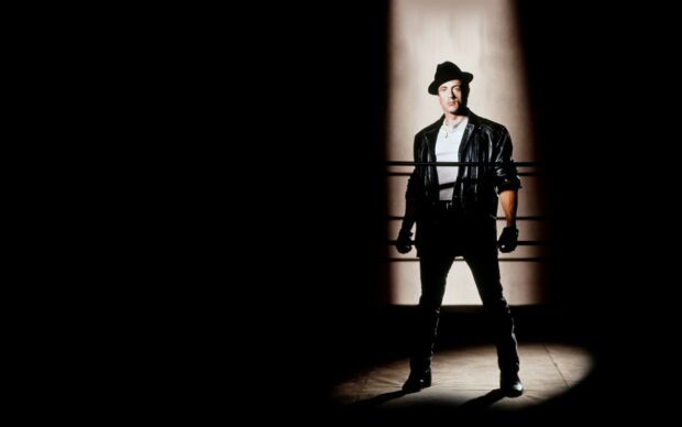 Rocky wearing a leather jacket and hat standing confidently in a boxing ring spotlight