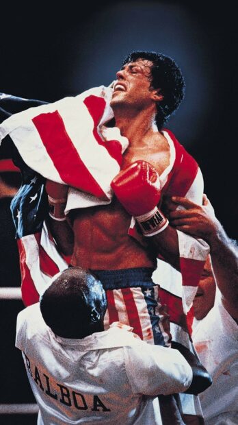 Rocky celebrating victory wrapped in American flag after boxing match with trainer holding him up