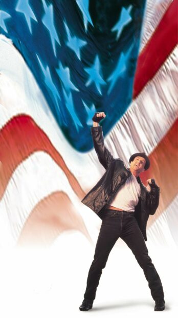 A man in a leather jacket and hat celebrating confidently with a raised fist in front of a large American flag