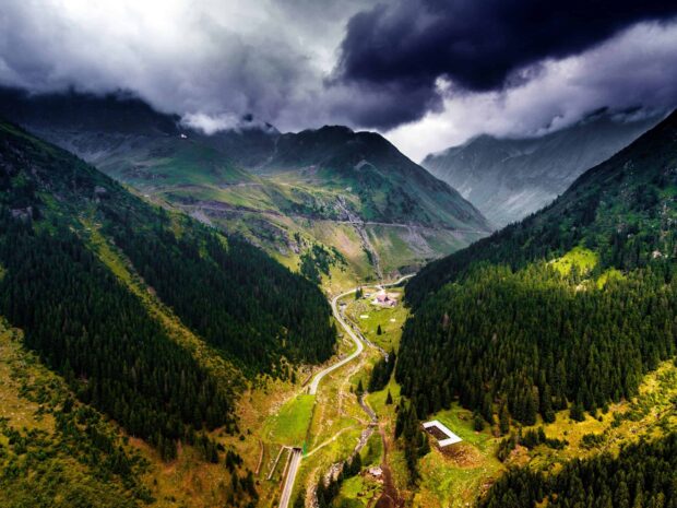 A winding road through lush green mountains in Romania surrounded by dense forests under dramatic clouds
