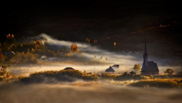 A misty Romanian village with autumn trees and a traditional church surrounded by fog in the morning