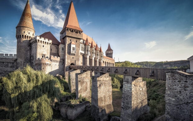 Historic Romania castle with stone bridge and lush green trees under blue sky