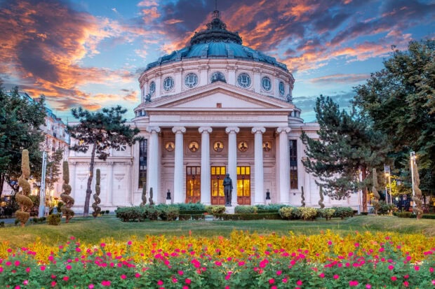 The Romanian Athenaeum surrounded by vibrant flowers and trees under a colorful sunset sky