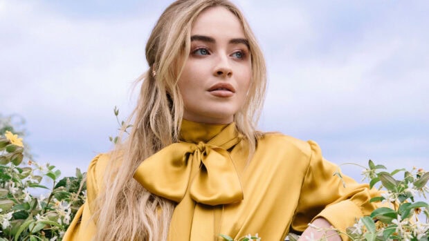 Sabrina Carpenter wearing a golden yellow blouse standing among green plants
