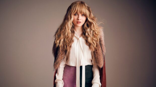 Sabrina Carpenter wearing a white blouse and a fur coat standing against a plain background