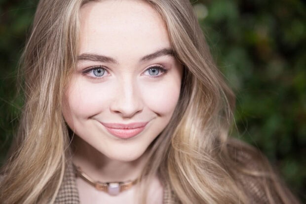 Sabrina Carpenter with long blonde hair smiling softly in a natural outdoor setting