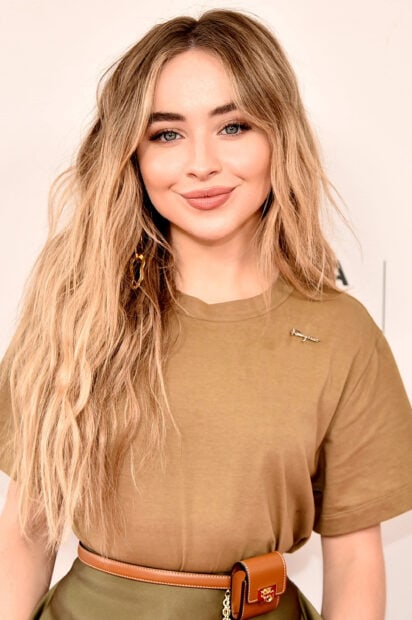 Sabrina Carpenter wearing a brown shirt and a belt smiling with long blonde hair