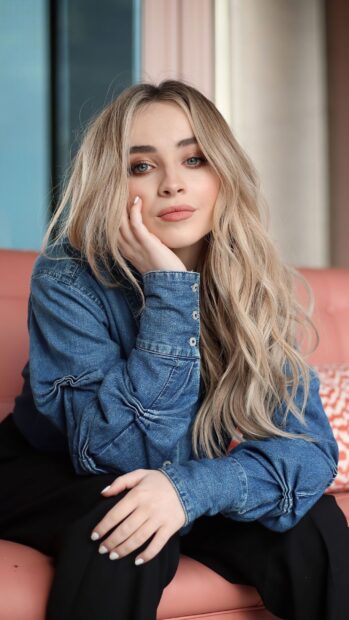 Sabrina Carpenter posing with her hand on her face wearing a denim jacket and black pants