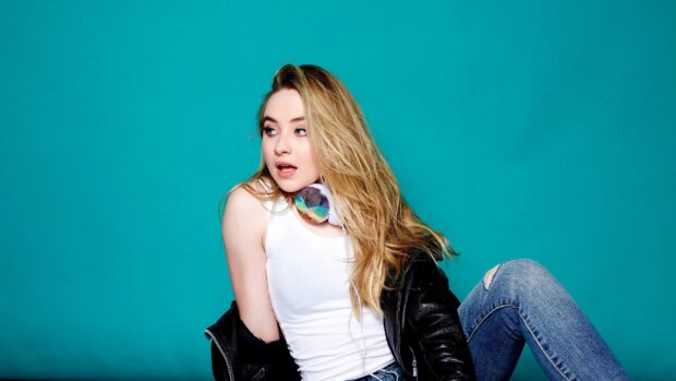 Sabrina Carpenter sitting against a teal background wearing headphones and a leather jacket