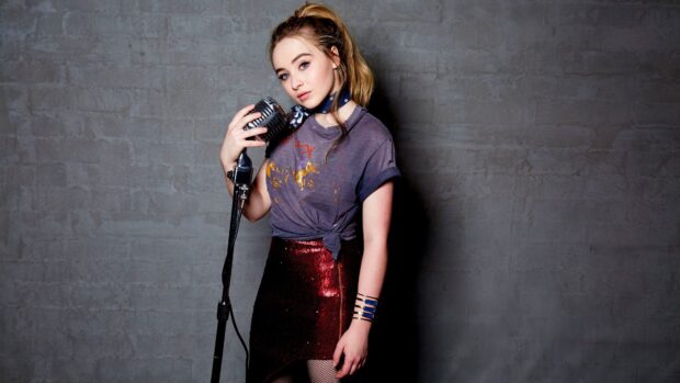 Sabrina Carpenter standing with a vintage microphone in front of a gray brick wall