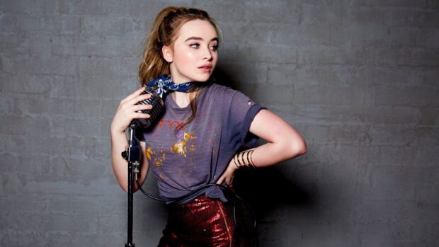Sabrina Carpenter wearing a vintage style shirt standing with a microphone against a gray wall