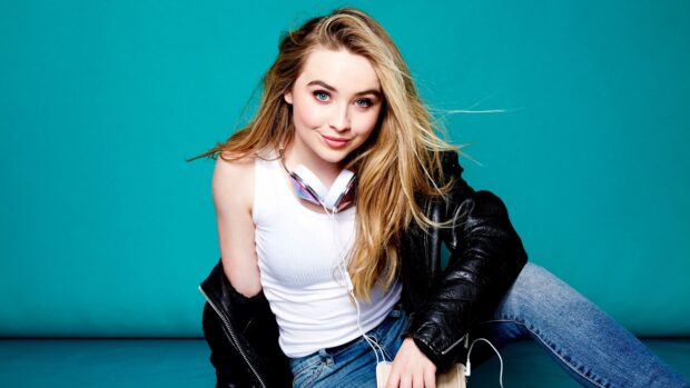 Sabrina Carpenter wearing a white tank top and leather jacket sitting against a blue background