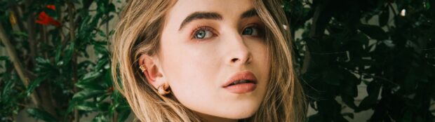 Sabrina Carpenter with blonde hair and blue eyes wearing gold earrings in a natural outdoor setting