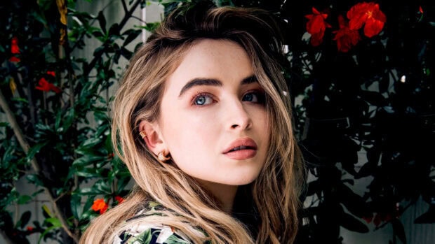 Sabrina Carpenter with natural makeup and floral outfit standing near green leaves and red flowers
