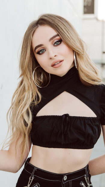 Sabrina Carpenter posing in a black top with long blonde hair and hoop earrings