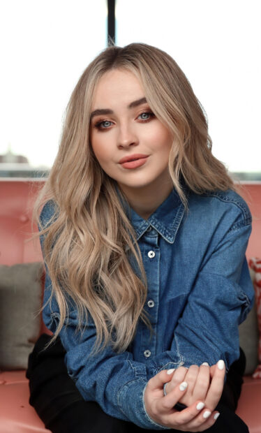 Sabrina Carpenter wearing a denim shirt and sitting on a sofa looking at the camera