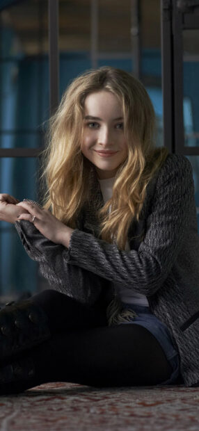 Sabrina Carpenter sitting indoors with a smile and long blonde hair
