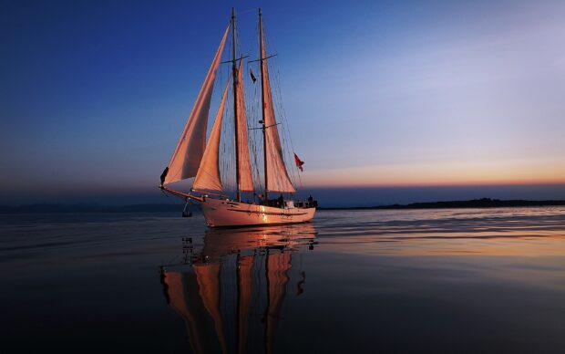 White sail boat sailing at sunset on calm water with clear sky and reflections