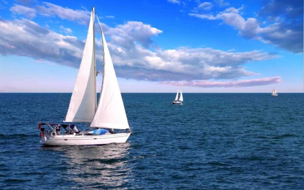A sail boat gliding across the ocean with white sails under a blue sky with scattered clouds