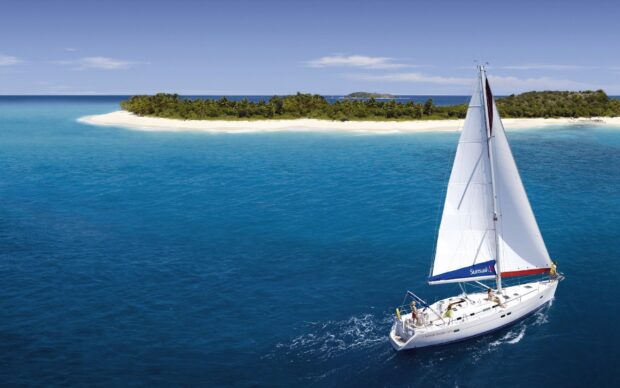 A sail boat cruising on clear blue ocean near a tropical island with white sandy beach