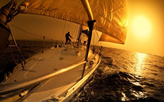 A sail boat sailing on the sea during a golden sunset with a person on deck