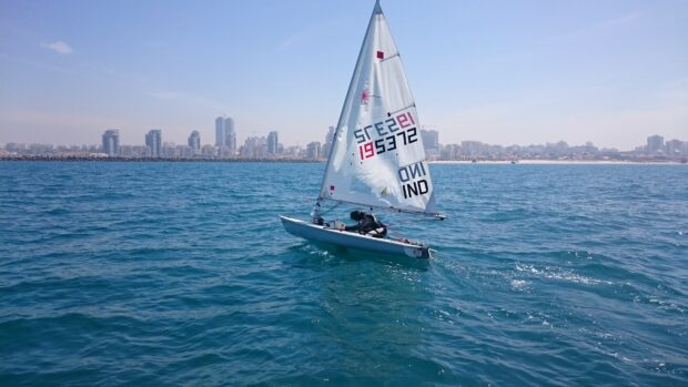 A sail boat navigating clear blue waters near a coastal city skyline
