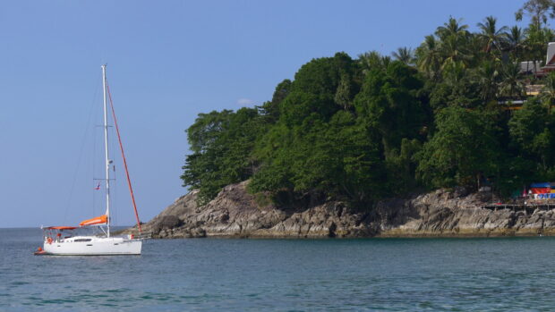 A sail boat near a rocky island covered with green trees in clear water