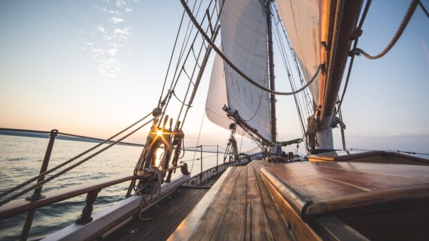 A sail boat with white sails gliding on calm water during sunset