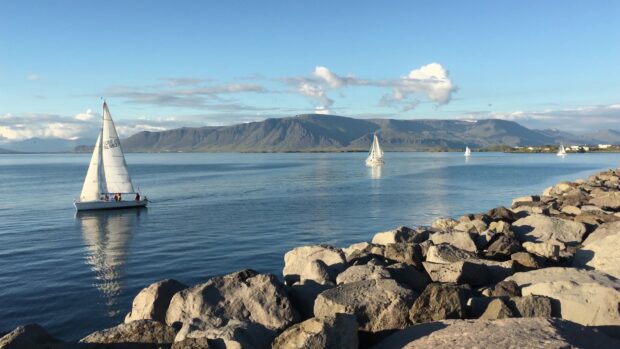 Sail boat sailing on calm water near rocky shore with mountains in the background