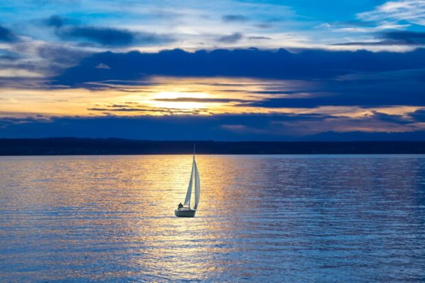 Sail boat sailing on calm water during sunset with dramatic sky and distant horizon