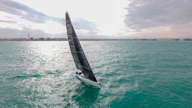 A sail boat gliding across the ocean near the coastline under a cloudy sky