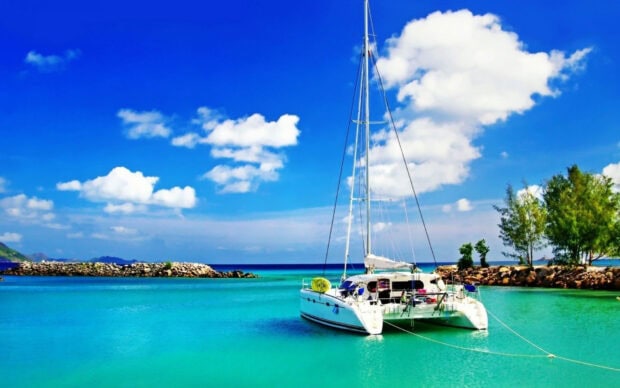 A sail boat anchored in clear blue water near a rocky shore under a bright sky