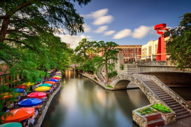 Colorful umbrellas along the San Antonio Texas Riverwalk with a stone bridge and city buildings in the background